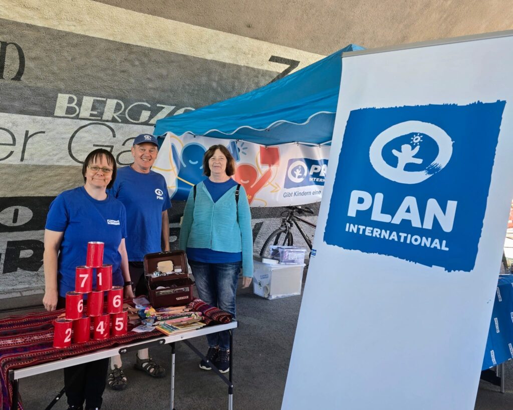 Drei Menschen stehen lächelnd an einem Stand von Plan International, der mit einem blauen Vordach, Informationsmaterialien, einem Stapel nummerierter roter Becher und einem großen Plan-International-Banner vor einer Wand mit Wandmalerei ausgestattet ist.